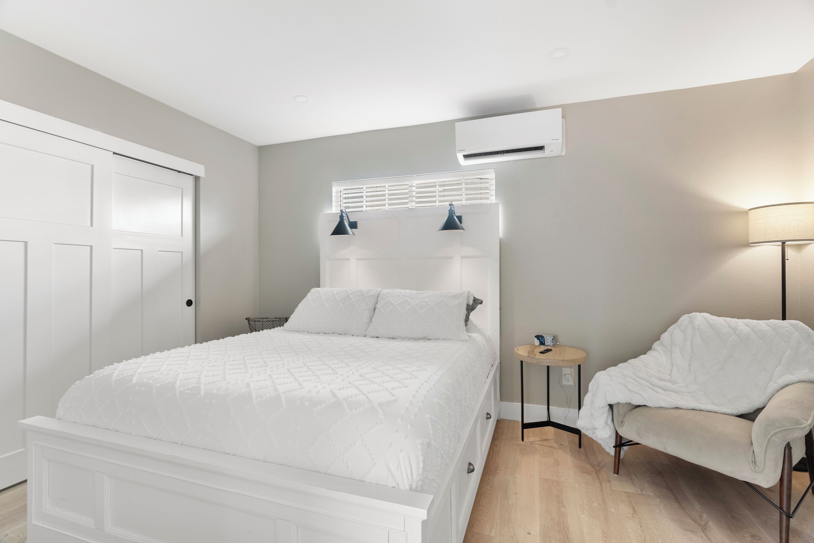 A bedroom with a bed, bedside table, chair, and an air conditioning unit.