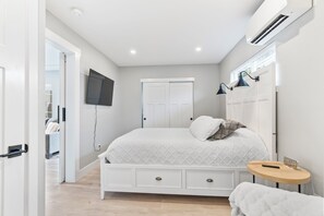 A bedroom with a bed, a bedside table, a TV mounted on the wall, a wardrobe, and a door leading to another room. - Petite Perfection | Grason Properties (Puyallup)