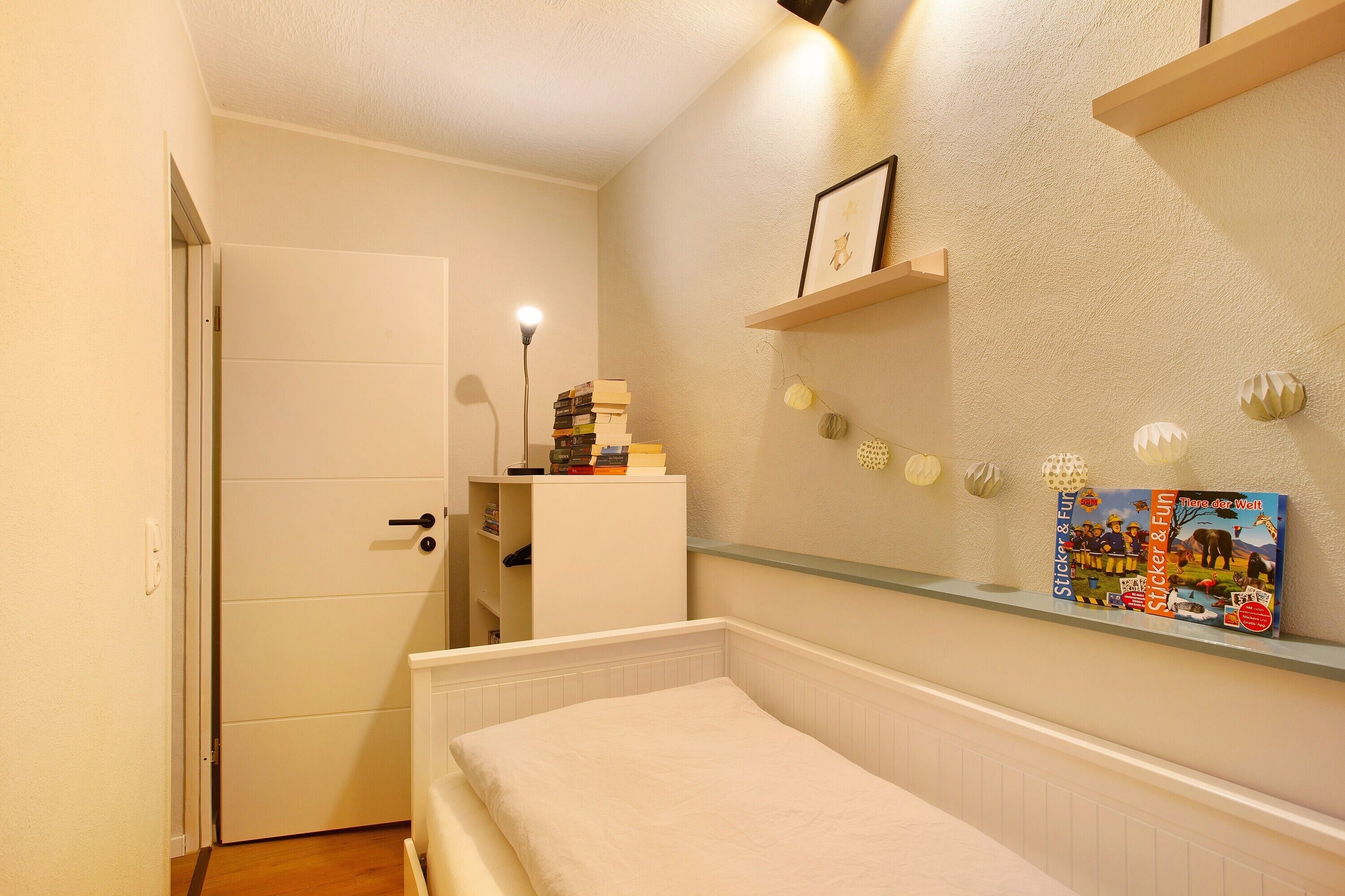 Ein Schlafzimmer mit einem Bett, einem weißen Kleiderschrank, einem Regal mit Büchern und einem gerahmten Bild sowie einer Tür.