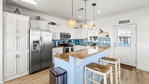 Une cuisine moderne avec des meubles blancs, des appareils en acier inoxydable, un îlot central doté d’un comptoir en granit et des luminaires suspendus.