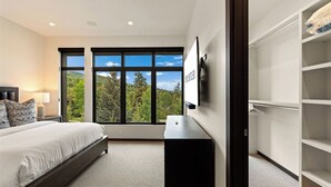 Een slaapkamer met een bed, een televisie, een kledingkast en een groot raam met uitzicht op bomen en heuvels.