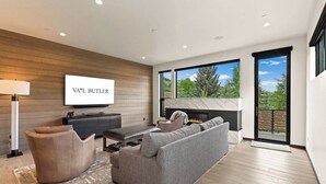Een moderne woonkamer met een flatscreen televisie, een open haard en een groot raam met uitzicht op bomen.