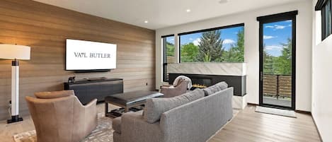 A modern living room with a flat-screen TV, a fireplace, and a large window offering a view of trees.