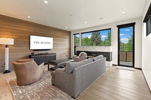 A modern living room with a flat-screen TV, a fireplace, and a large window offering a view of trees. - Modern Condo minutes from Beaver Creek! Basecamp14 (Avon)