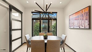Een moderne eethoek met een ronde tafel, zes stoelen, een centrale lamp en een groot raam met uitzicht op gebouwen en bomen.