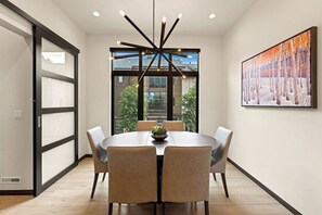 A modern dining area with a round table, six chairs, a central light fixture, and a large window with a view of buildings and trees. - Modern Condo minutes from Beaver Creek! Basecamp14 (Avon)