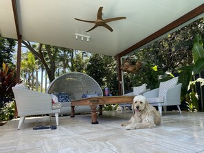Uma área de estar coberta ao ar livre, com ventoinha no teto, mesa de madeira e um cão.