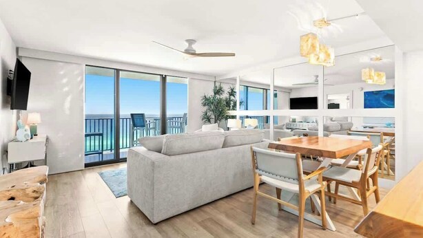 A modern living room with a grey sofa, wooden dining table, and chairs. There is a large window with a view of the ocean.