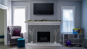 Un salon avec une cheminée, un fauteuil gris, un pouf bleu et un téléviseur fixé au mur.