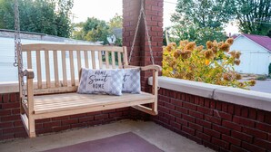 Un banc-balançoire en bois avec deux coussins, dont l’un porte l’inscription « HOME Sweet HOME », adossé à un mur de briques. On aperçoit une petite plante et un bâtiment blanc en arrière-plan.