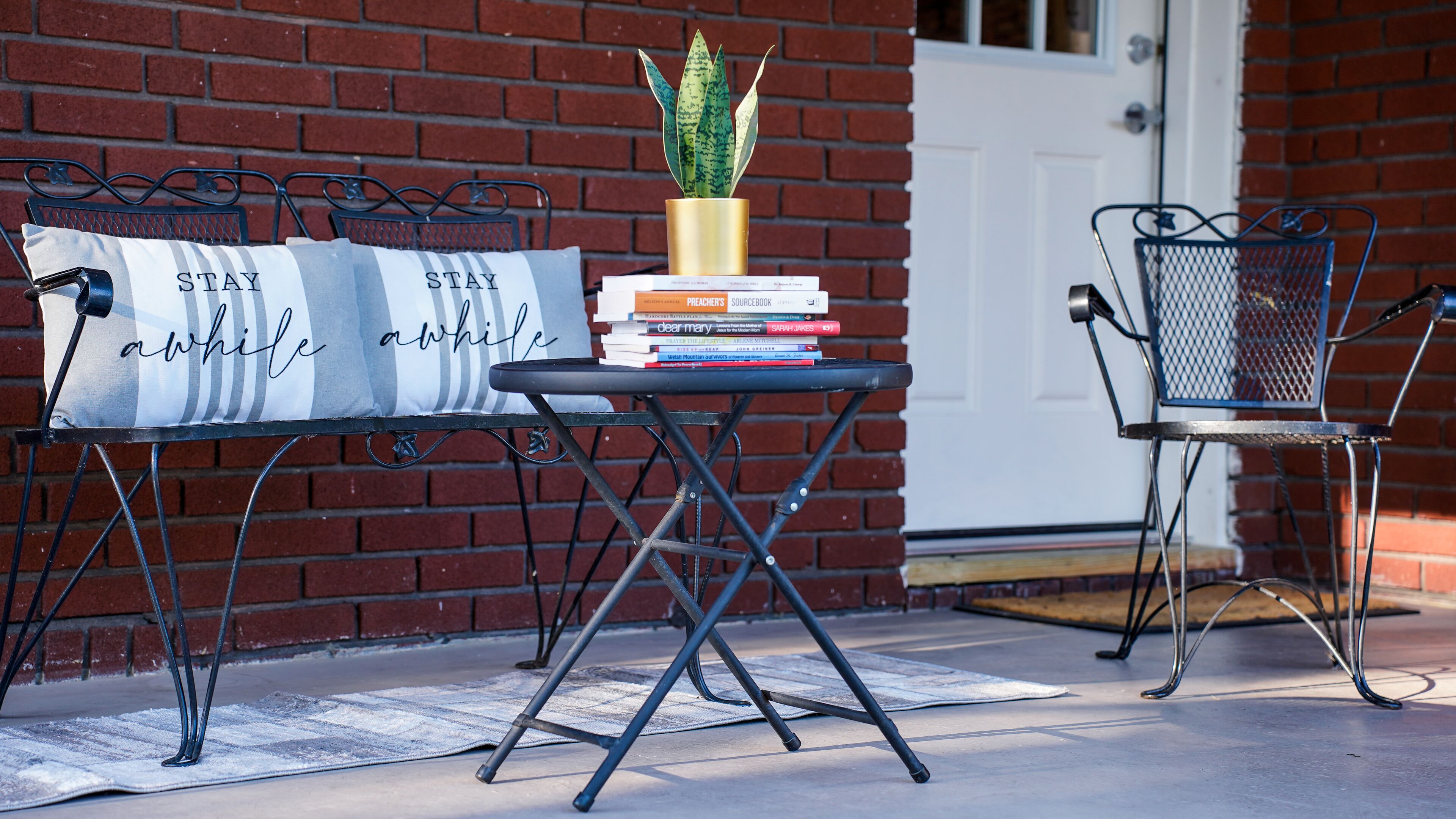 A metal table with a stack of books and a potted plant, a bench with a cushion, and a metal chair against a brick wall.