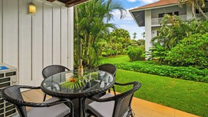 A glass-top table with wicker chairs, a bottle, and a glass on it, set against a white wall with a window overlooking a lush garden and a building with balconies. - Kiahuna Paradise @ Poipu with 2  AC units 
Family size condo with patio (Koloa)