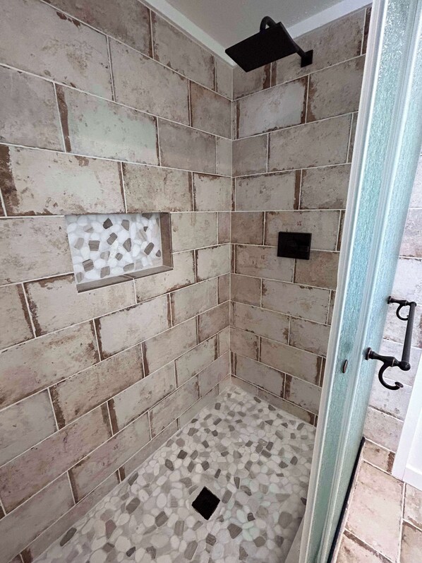 A shower area with a tiled wall and floor, a black showerhead, and a small window. - Mucho Gusto PLUS Casita: 3br 3ba, 2.4 miles from BBNP (Terlingua)