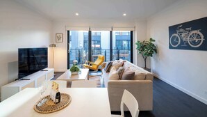 A modern living room with a sofa, a dining table, a TV, and a balcony with a view of buildings. - Bright 3BR 3 Level Townhouse Port Melbourne (Port Melbourne)