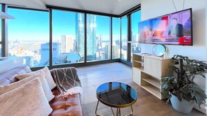 A modern living room with a leather sofa, a glass coffee table, a TV mounted on the wall, and a large window offering a city view. - LV42 High-Rise Condo CBD Panoramic City View (Melbourne)