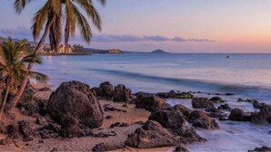 Une plage avec un rivage rocheux, des palmiers et une mer calme au coucher du soleil.