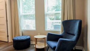 Ein Zimmer mit einem dunkelblauen Sessel, einem kleinen runden Holztisch und passendem Hocker. Zwei Fenster mit Jalousien sind vorhanden, und eine Tür ist links zu sehen.