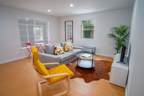Uma sala de estar moderna com um sofá cinzento, uma poltrona amarela, uma mesa de centro de vidro e um suporte branco para TV.