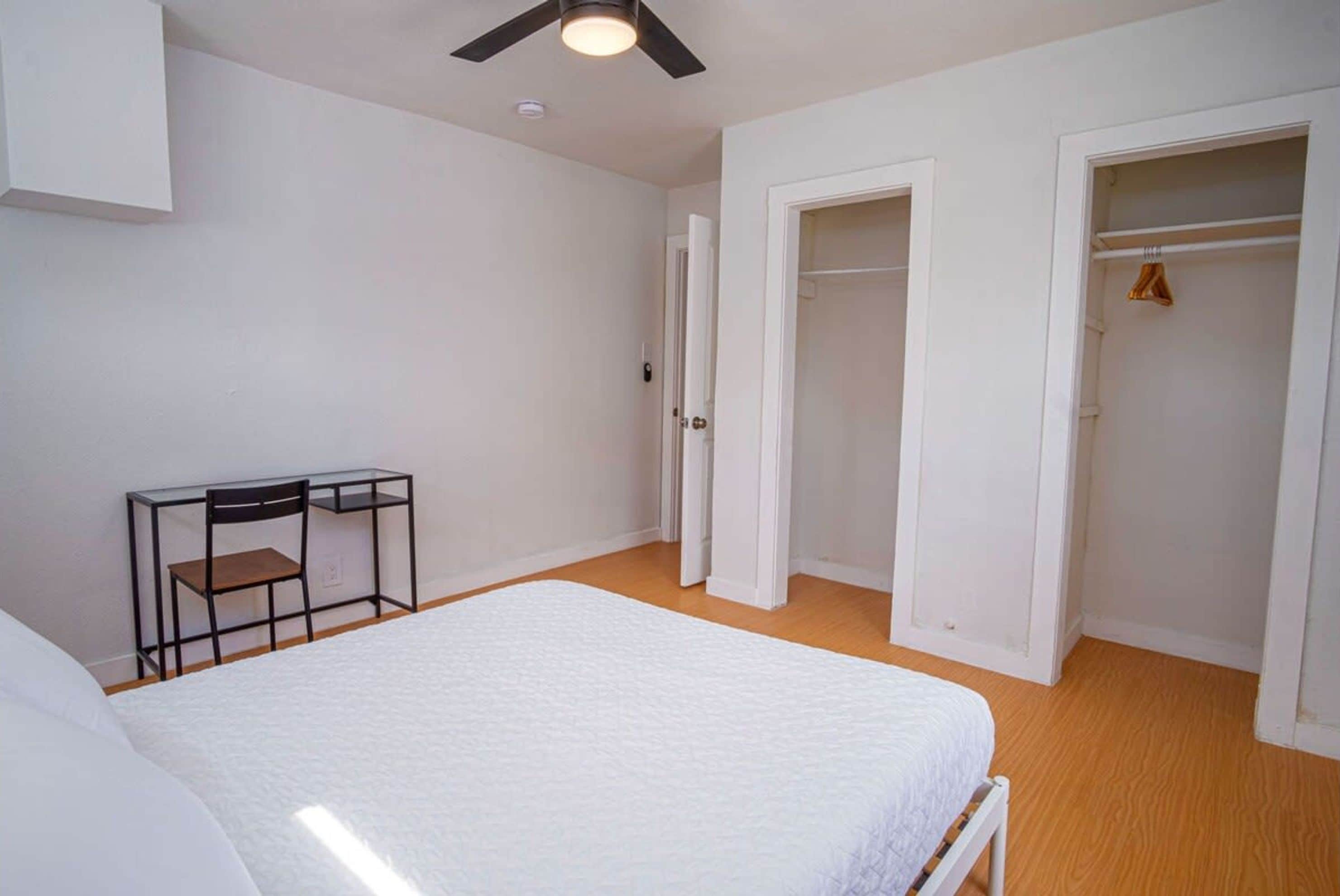 A bedroom with a bed, a desk, and a chair. There are two doors leading to closets.