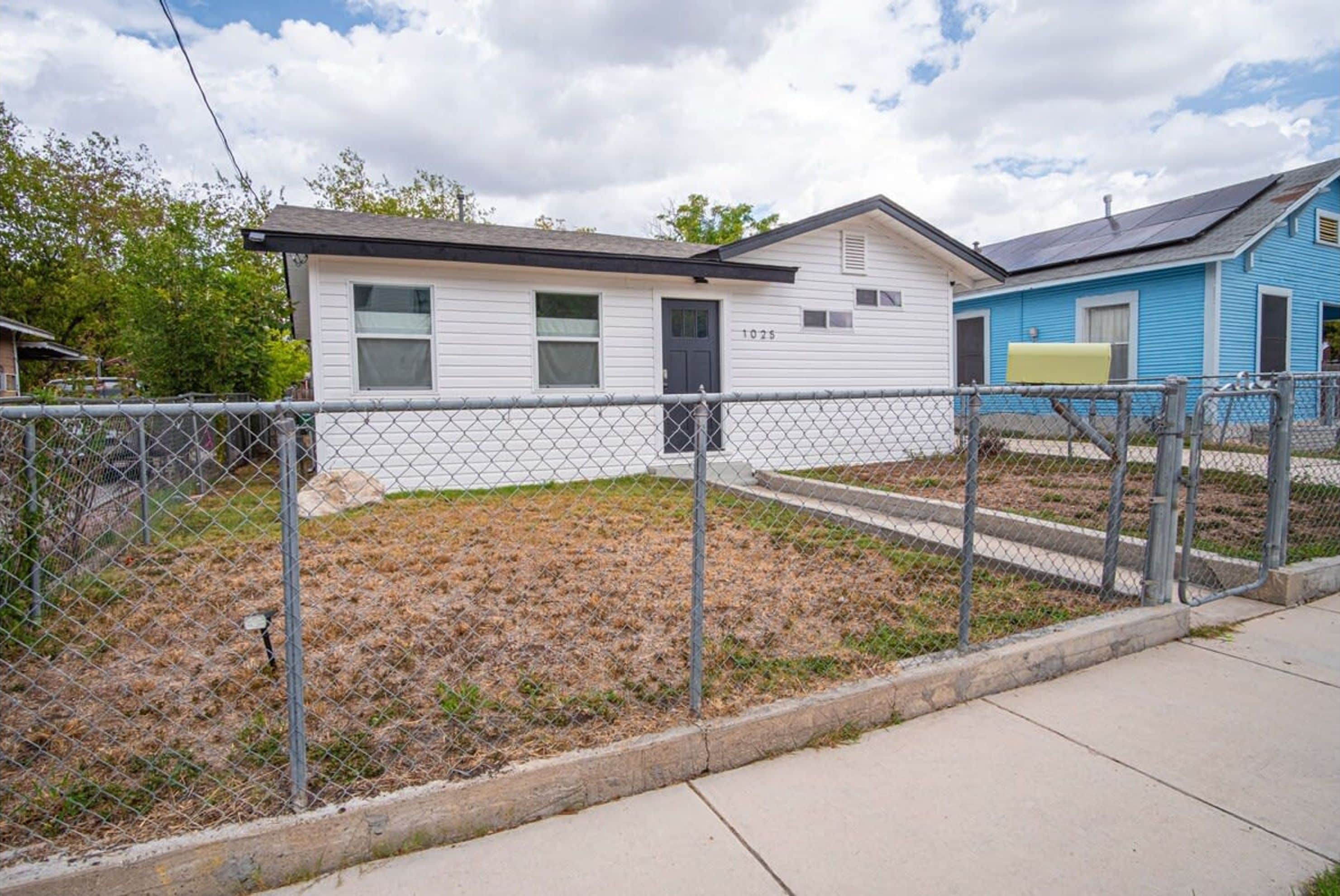 A single-story house with a black door and a mailbox, surrounded by a chain-link fence.