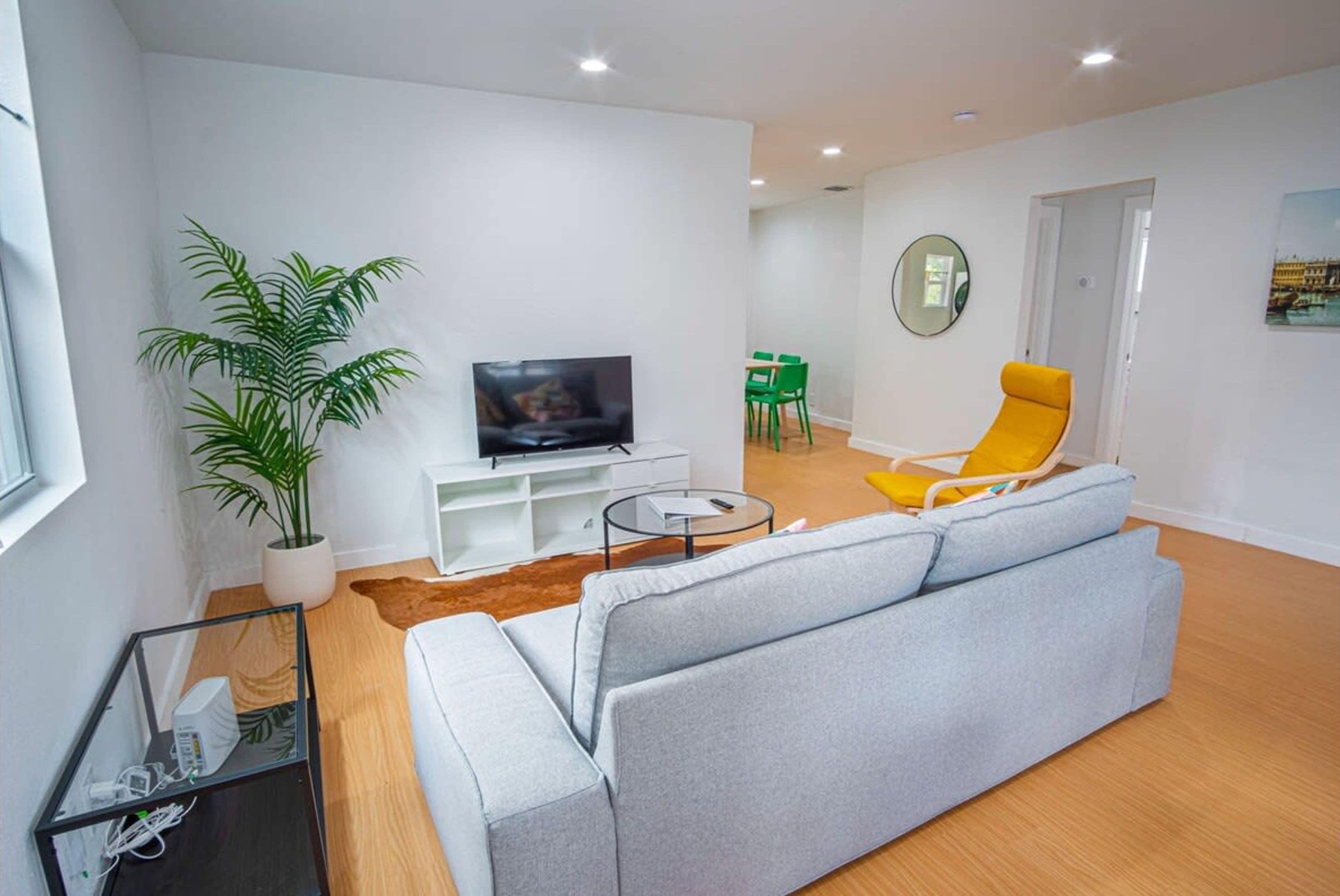 Ein modernes Wohnzimmer mit grauem Sofa, einem gelben Sessel, einem Fernseher auf einem weißen Schrank, einer Zimmerpflanze und einem Essbereich im Hintergrund.