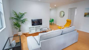 Ein modernes Wohnzimmer mit grauem Sofa, einem gelben Sessel, einem Fernseher auf einem weißen Schrank, einer Zimmerpflanze und einem Essbereich im Hintergrund.