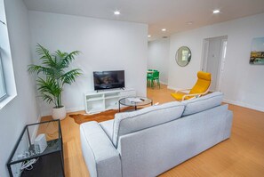 A modern living room with a grey sofa, a yellow armchair, a TV on a white stand, a potted plant, and a dining area in the background. - Serene 2BR 1BA Residence in Peaceful San Antonio (San Antonio)