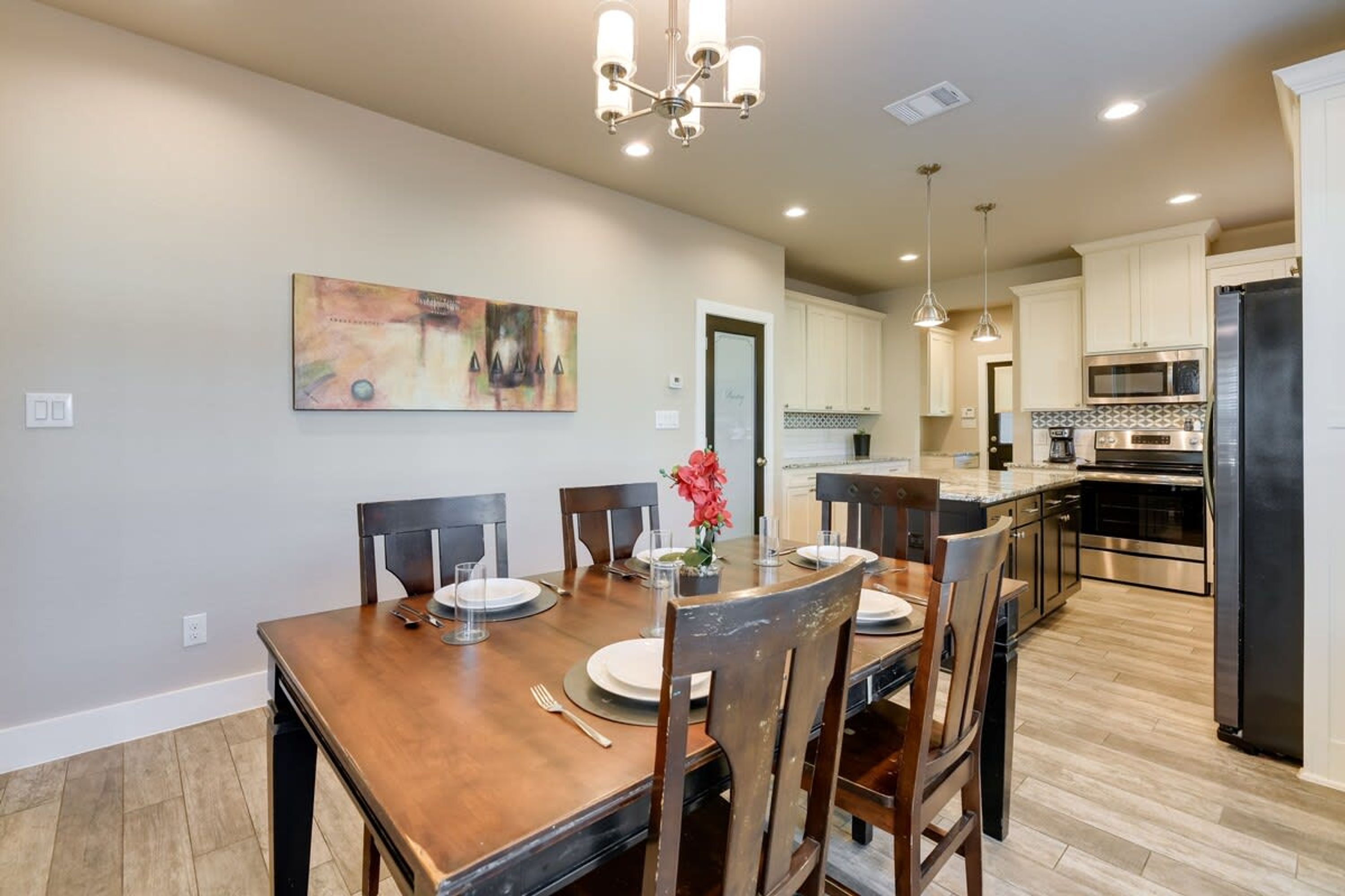A dining area with a wooden table set for four, a chandelier, and a kitchen in the background.