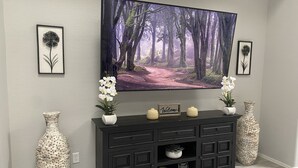 A modern living room with a large flat-screen TV mounted on the wall, a dark wooden console table with decorative items, and two large woven vases. - Super close to all the fun, short walk to Westgate Entertainment District.   (Glendale)