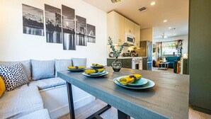 A modern kitchen with a dining area, featuring a grey sofa, a wooden dining table with plates and napkins, and cityscape artwork on the wall.
