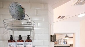A bathroom with a white tiled wall, a shower area, and a sink with a faucet. There are three bottles of Nectar shampoo on a shelf.