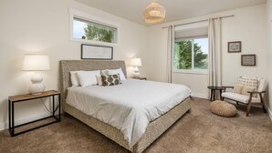 Un dormitorio con una cama grande, dos mesitas de noche con lámparas, una lámpara colgante, una ventana con vistas a árboles y una silla con cojín.
