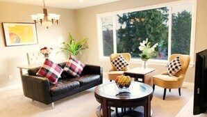 Ein Wohnzimmer mit einer Couch, zwei Sesseln, einem Couchtisch und einem Fernseher. Es gibt ein Fenster mit Blick auf Bäume und eine Kristallleuchter.