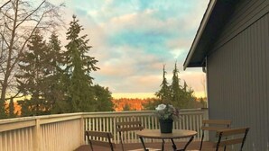 Eine Holzterrasse mit rundem Tisch und Stühlen, umgeben von einem weißen Zaun, mit Blick auf eine bewaldete Landschaft.