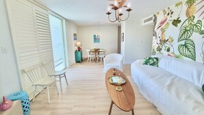 A living room with a white sofa, a wooden coffee table, a dining area with a table and chairs, and a wall with a tropical plant print.
