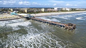 A pier extending into the ocean with buildings and a beach in the background.