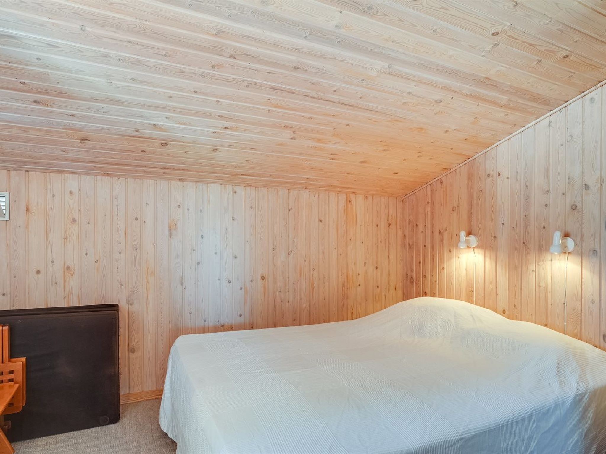 A room with wooden walls and ceiling, a bed with a white bedspread, a small table, and two wall-mounted lights.