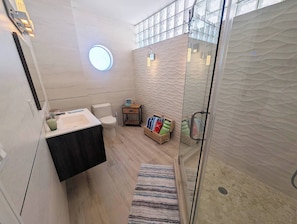 A modern bathroom with a glass shower enclosure, a toilet, a sink with a dark countertop, and a small bedside table with towels.