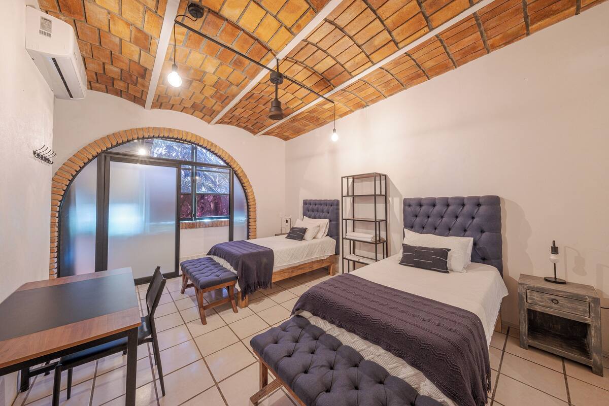 A bedroom with a brick ceiling, two beds, a dining table, and a bench.