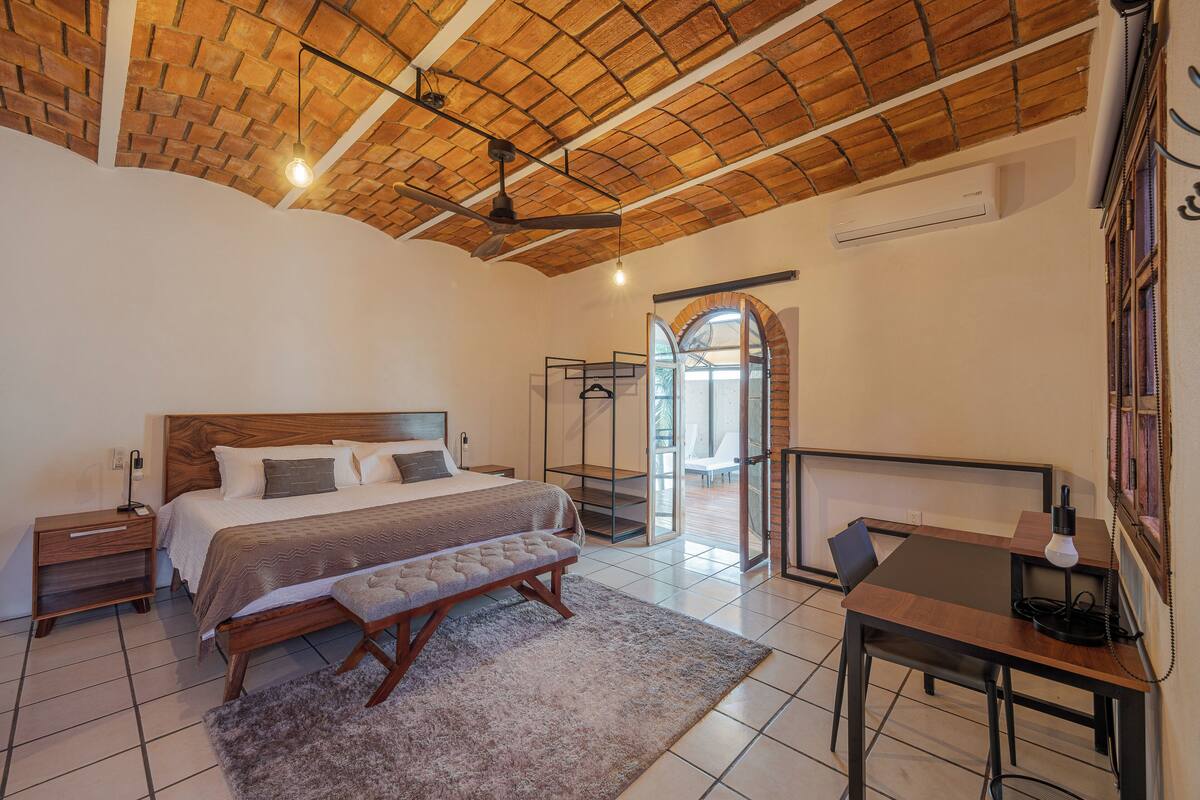 A bedroom with a wooden ceiling, a large bed, a bench, a desk, and a chair.