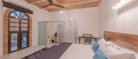 Quarto com cama grande, ventilador de teto, janela e porta de acesso a um banheiro.