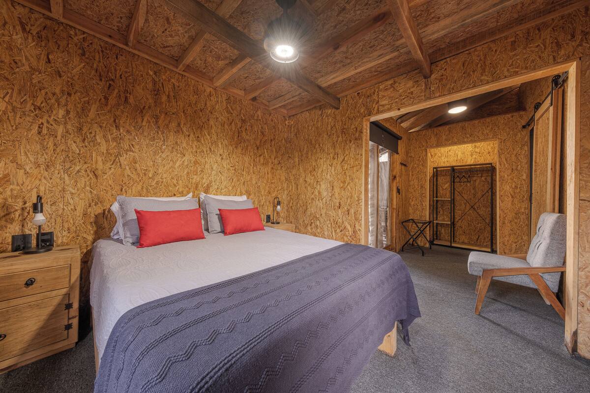 A wooden-paneled room with a bed, a nightstand, a chair, and a door leading to another room.