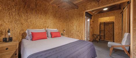 A wooden-paneled room with a bed, a nightstand, a chair, and a door leading to another room.