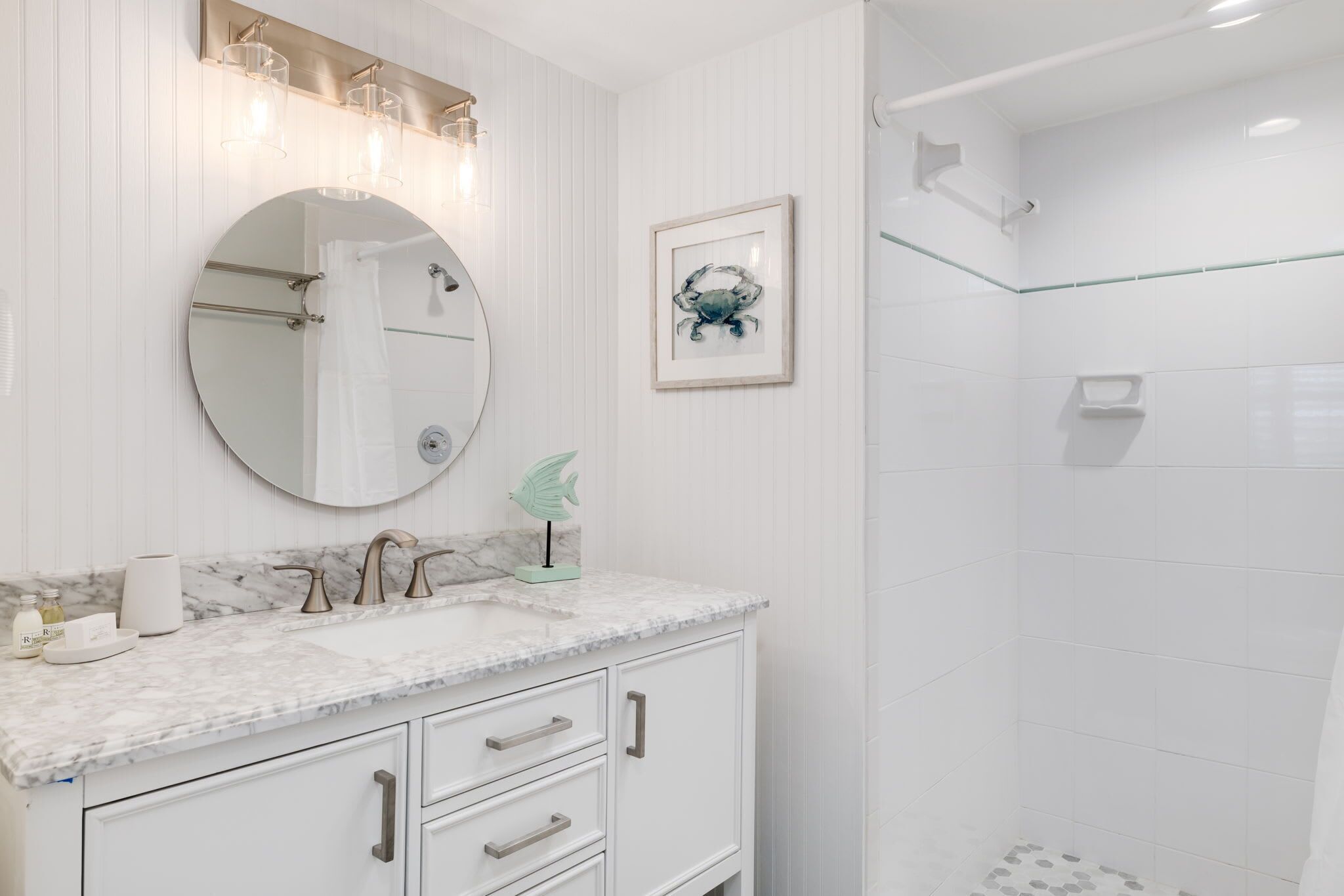 A bathroom with a marble vanity, a round mirror, a framed crab artwork, and a glass shower enclosure.