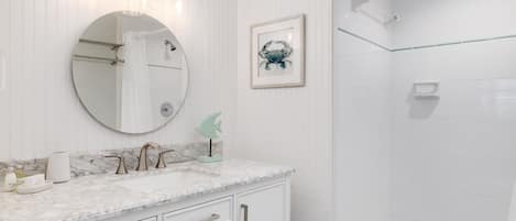 A bathroom with a marble vanity, a round mirror, a framed crab artwork, and a glass shower enclosure.