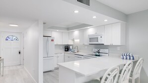 A modern kitchen with white cabinetry, a central island, and a dining area with white chairs. - Breakers West C2 | Stunning Top Floor Beach Residence | Steps to the Sand (Sanibel)