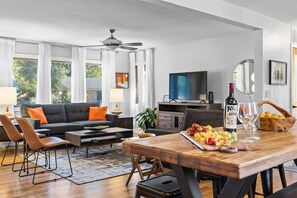 Una sala de estar moderna con un sofá, sillones, una mesa de centro, una mesa de comedor con comida y bebidas, un televisor y un ventilador de techo.