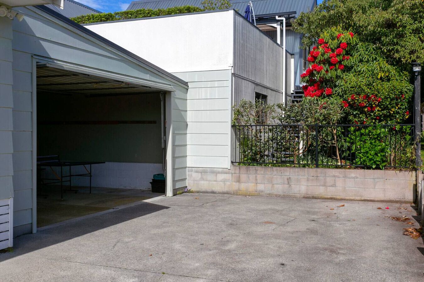Un garage avec une table et des chaises à l’intérieur, une allée en béton et un arbuste aux fleurs rouges.