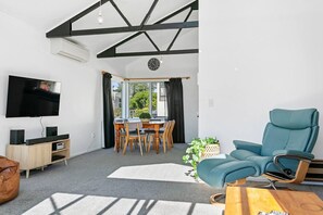A modern living room with a flat-screen TV, wooden furniture, a teal armchair, and a dining area with natural light.