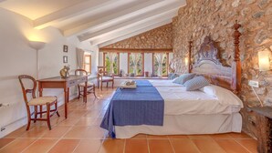 Ein Schlafzimmer mit einer Steinsäule, einem Bett mit Holz-Kopfteil, einem Schreibtisch mit Stühlen und Blick durch große Fenster.